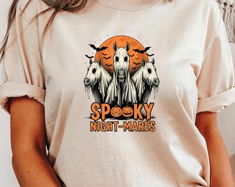 May include: Beige t-shirt featuring a Halloween design. The graphic includes three ghostly horses in front of an orange moon, with bats and the text "Spooky Night-mares". The design is in orange and white.
