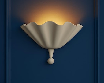 May include: A cream-colored wall sconce with a scalloped, fan-like shade. The sconce is mounted on a dark blue wall, with a warm light emanating from within the shade. The fixture has a small, round decorative element at the bottom.