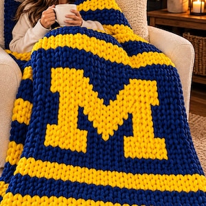 May include: A chunky knit throw blanket featuring a bold, repeating striped pattern in deep blue and bright yellow. The center of the blanket displays a large, block-style letter M in yellow, integrated into the thick, textured yarn weave.