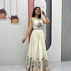 May include: A sleeveless, ivory-colored gown with intricate embroidery. The bodice features colorful floral and elephant motifs, while the skirt has a flowing, pleated design. The hemline is adorned with a detailed border, creating an elegant and festive look.
