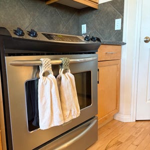 May include: A stainless steel oven with two white kitchen towels hanging from the handle. The towels are made of a textured fabric and are suspended by crocheted rings. The oven is in a kitchen with wooden cabinets and flooring.