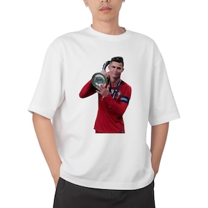 May include: White oversized t-shirt featuring a graphic of a soccer player in a red jersey holding a trophy. The player is smiling. The shirt is made of a soft, comfortable material.