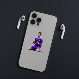May include: A silver smartphone case featuring an image of a soccer player in a purple uniform, celebrating on his knees. Two white wireless earbuds are positioned on either side of the phone. The background is a dark gray surface.