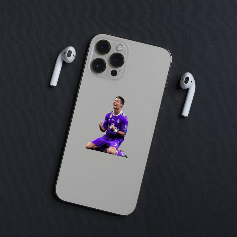 May include: A silver smartphone case featuring an image of a soccer player in a purple uniform, celebrating on his knees. Two white wireless earbuds are positioned on either side of the phone. The background is a dark gray surface.