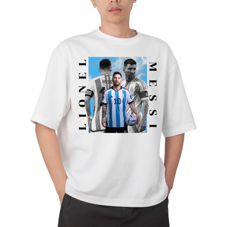 Puede incluir: Camiseta blanca con un gr&aacute;fico de Lionel Messi con una camiseta de f&uacute;tbol azul y blanca, sosteniendo un bal&oacute;n de f&uacute;tbol. Las palabras "MESSI" y "LIONEL" est&aacute;n impresas verticalmente en la camiseta.