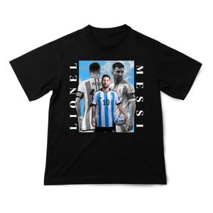 Puede incluir: Camiseta negra con un gr&aacute;fico de Lionel Messi con su uniforme de f&uacute;tbol argentino, con el texto "Lionel Messi" en letras verticales blancas. La imagen muestra a Messi sosteniendo un bal&oacute;n de f&uacute;tbol.