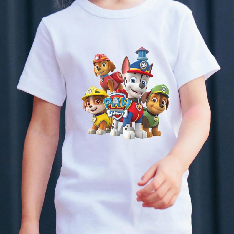 May include: A white short-sleeved t-shirt featuring a colorful graphic print of four animated dog characters from Paw Patrol. The design includes the Paw Patrol logo in the center, with the characters wearing various uniforms in red, yellow, and green.