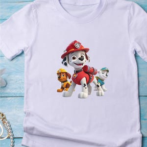 May include: A white short-sleeved t-shirt featuring a graphic print of three cartoon dogs from Paw Patrol. The central character is a Dalmatian wearing a red firefighter helmet and vest, flanked by a brown dog in a yellow hat and a white dog.