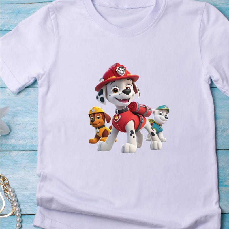 May include: A white short-sleeved t-shirt featuring a graphic print of three cartoon dogs from Paw Patrol. The central character is a Dalmatian wearing a red firefighter helmet and vest, flanked by a brown dog in a yellow hat and a white dog.