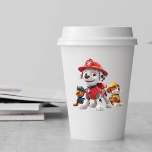 May include: A white reusable travel cup featuring a graphic print of three cartoon dogs from Paw Patrol. The central character is a white and black Dalmatian wearing a red firefighter hat and vest, flanked by a blue puppy and a yellow puppy.