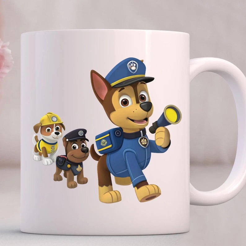 May include: A white ceramic mug featuring a colorful graphic of three cartoon dogs from Paw Patrol. The largest dog wears a blue police uniform and cap while holding a flashlight. Two smaller dogs in yellow and blue uniforms stand behind it.