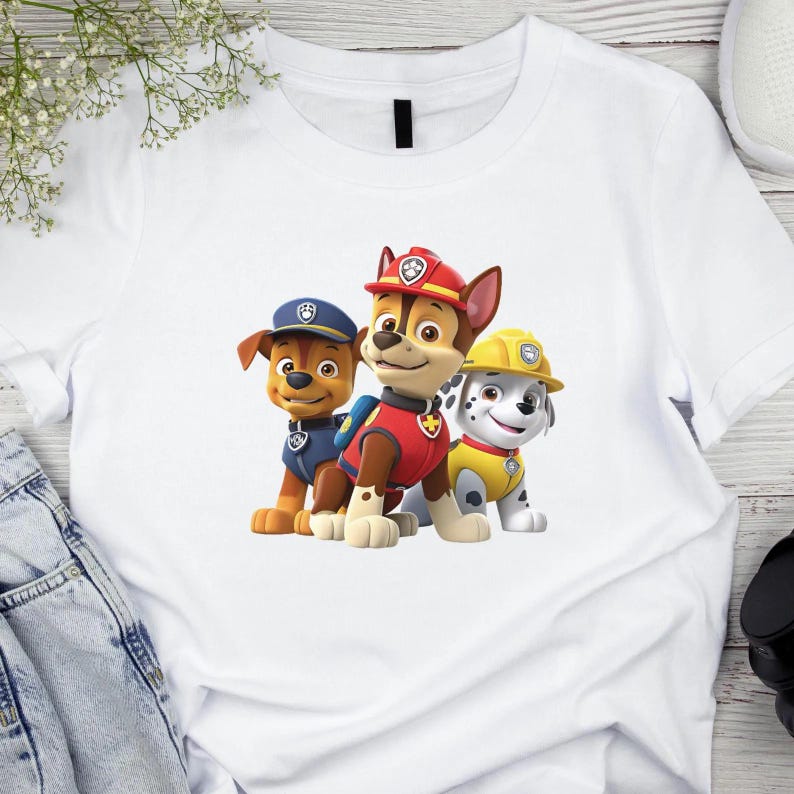 May include: A white short-sleeved t-shirt featuring a graphic print of three cartoon dogs from Paw Patrol. The characters include a brown dog in a blue uniform, a brown dog in a red uniform, and a white spotted dog in a yellow construction hat.