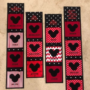 May include: A black and red fabric wall organizer with Mickey Mouse ears and names for each pocket. The names include: The Hawkins Family, Mom & Dad, Cole, Evan, Drew, The Choi Family, Mom & Dad, The Agena Family, Mom, Ashlee, Colby, Josie, Cruisin' with Mickey, and living the DREAM!, Eddie, Tiffany, Skyler, Kelsey.