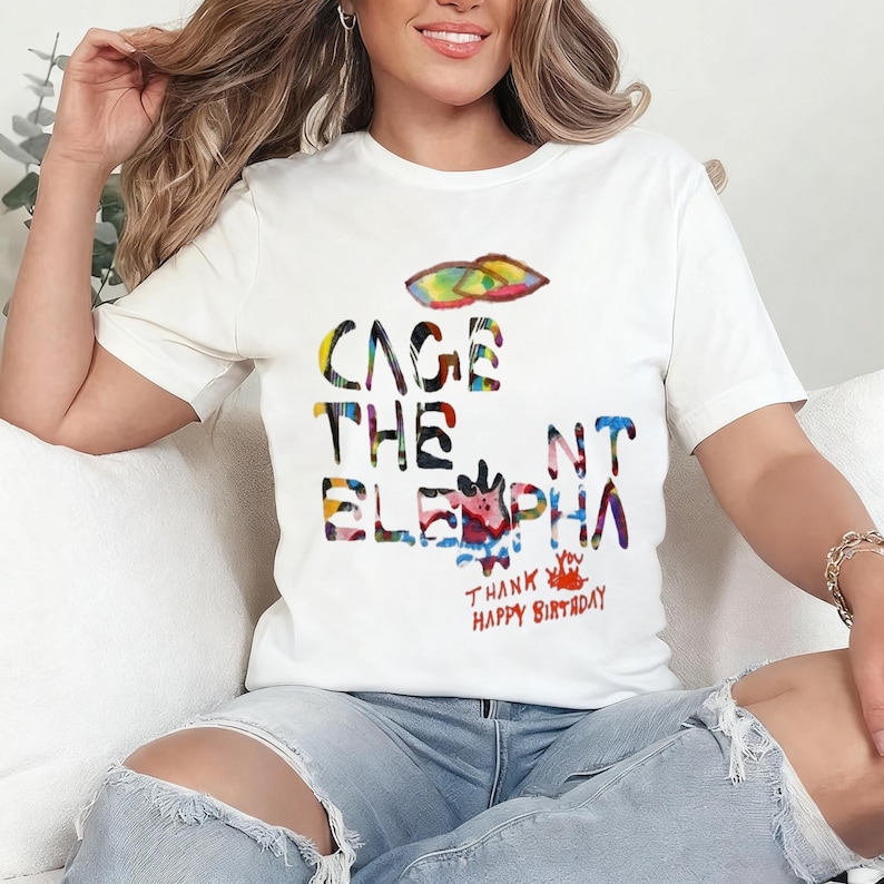 May include: White t-shirt with the text "CAGE THE ELEPHANT" in colorful, watercolor-style lettering. The shirt also features the text "THANK YOU HAPPY BIRTHDAY" in red. A colorful design is above the text.