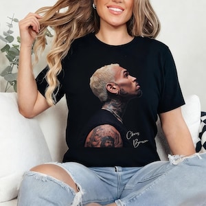 May include: Black t-shirt featuring a profile image of Chris Brown with a tattoo on the arm. The name "Chris Brown" is written in cursive script. The person in the image is wearing blue jeans.