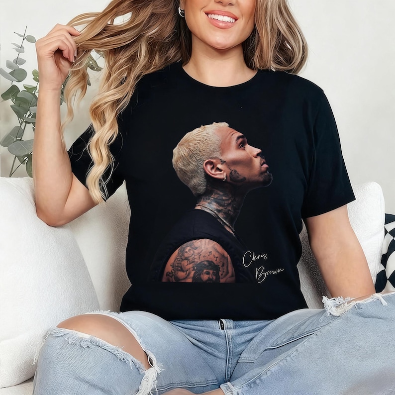 May include: Black t-shirt featuring a profile image of Chris Brown with a tattoo on the arm. The name "Chris Brown" is written in cursive script. The person in the image is wearing blue jeans.