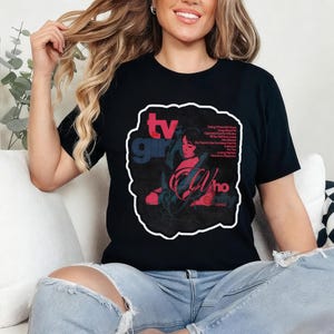 K&ouml;nnte beinhalten: Schwarzes T-Shirt mit einem grafischen Design. Das Design zeigt den Text "TV Girl" in Rot und Blau sowie "Who Really Cares" in einer stilisierten Schriftart. Ein Bild einer Frau in Rot und T&uuml;rkis ist ebenfalls enthalten.