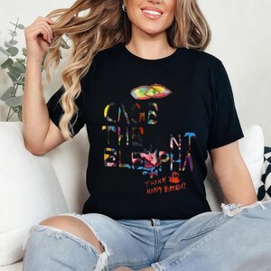 May include: Black t-shirt with a colorful graphic design. The design includes the words "CAGE THE ELEPHANT" in a vibrant, multi-colored font, along with the text "THANK HAPPY BIRTHDAY". The shirt is short-sleeved.