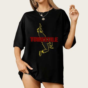 May include: Black t-shirt with the word "TURNSTILE" in red block letters and a yellow outline of a person in action. The shirt has short sleeves and a loose fit.