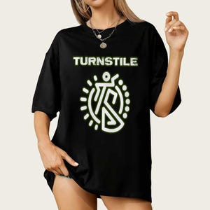 May include: Black t-shirt with the word "TURNSTILE" in a green, distressed font above a white graphic. The graphic is a stylized figure within a dotted circle. The shirt has short sleeves.