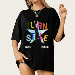 May include: Black t-shirt featuring the words "TURN STYLE" in colourful letters, a silhouette of a person, and the words "NEVER ENOUGH" in white. The design is centered on the shirt.