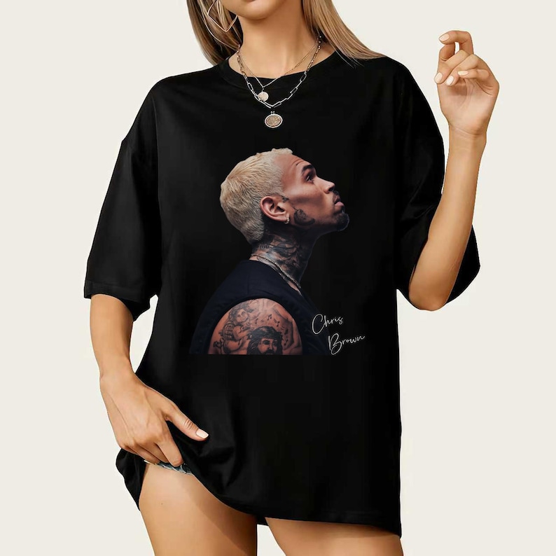 May include: Black t-shirt featuring a side profile image of Chris Brown with a tattoo on his arm and the text "Chris Brown" in a handwritten font. The image is printed on a black t-shirt.