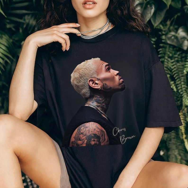 May include: Black t-shirt featuring a portrait of Chris Brown with blonde hair and tattoos. The image includes the artist's name in a handwritten style. The shirt is a casual style, suitable for everyday wear.