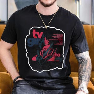 K&ouml;nnte beinhalten: Schwarzes T-Shirt mit einem grafischen Design mit dem Text "tv girl" in Blau und Rot und dem Wort "Who" in Rot. Das Design enth&auml;lt ein Portr&auml;t einer Frau und Songtitel.