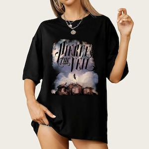 Puede incluir: Camiseta negra con el nombre de la banda "Pierce the Veil" en una fuente estilizada sobre una ilustración de una figura en un tejado. El gráfico tiene un fondo nublado. La camiseta tiene mangas cortas y un corte holgado.