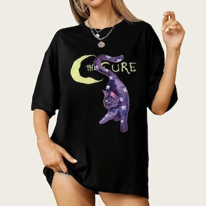以下が含まれることがあります： 白い星で飾られた紫色の猫のデザインが特徴の黒いTシャツ。三日月のようにアーチ型になっており、「THE CURE」の文字が書かれています。猫のデザインは黄色の三日月を背景にしています。