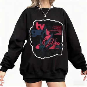 K&ouml;nnte beinhalten: Schwarzes Sweatshirt mit wei&szlig;em Kragen und Grafikdesign. Das Design zeigt die Worte "TV Girl" in Rot und Blau sowie eine Illustration einer Frau. Zus&auml;tzlicher Text ist sichtbar.