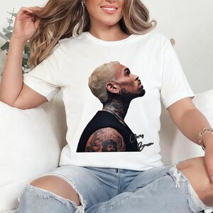 May include: White t-shirt featuring a portrait of Chris Brown in a black top, with the name "Chris Brown" in cursive script. The image is a side profile of the singer with tattoos visible on his arm and neck.
