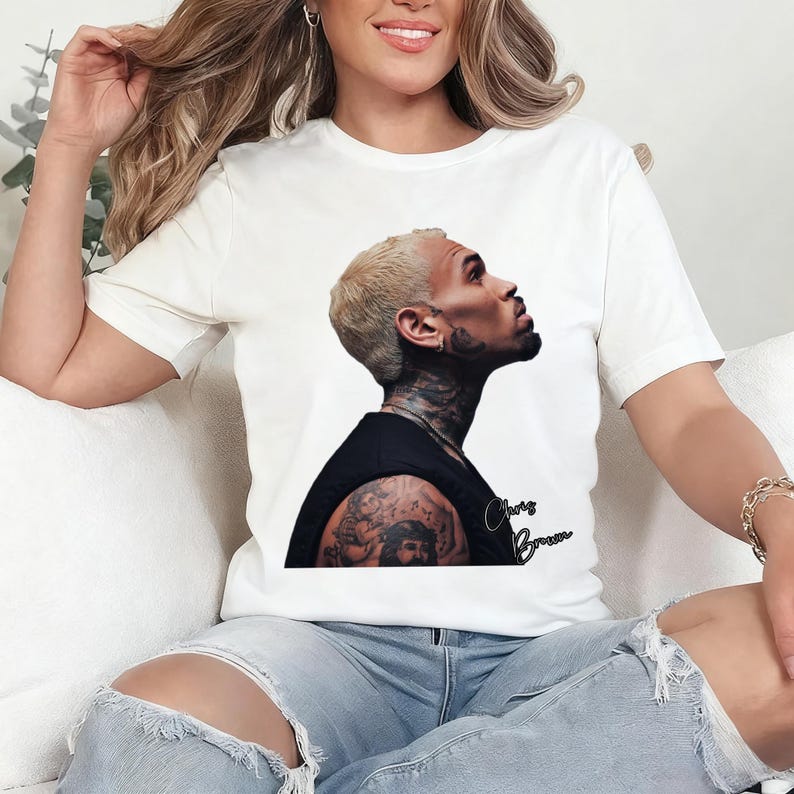 May include: White t-shirt featuring a portrait of Chris Brown in a black top, with the name "Chris Brown" in cursive script. The image is a side profile of the singer with tattoos visible on his arm and neck.