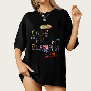 Op de afbeelding: Zwart T-shirt met de tekst "CAGE THE ELEPHANT" in kleurrijke, gestileerde letters. Het shirt bevat ook de zinnen "THANK HAPPY BIRTHDAY" in een levendig, veelkleurig ontwerp. Een afbeelding van een kleurrijke lip staat boven de tekst.
