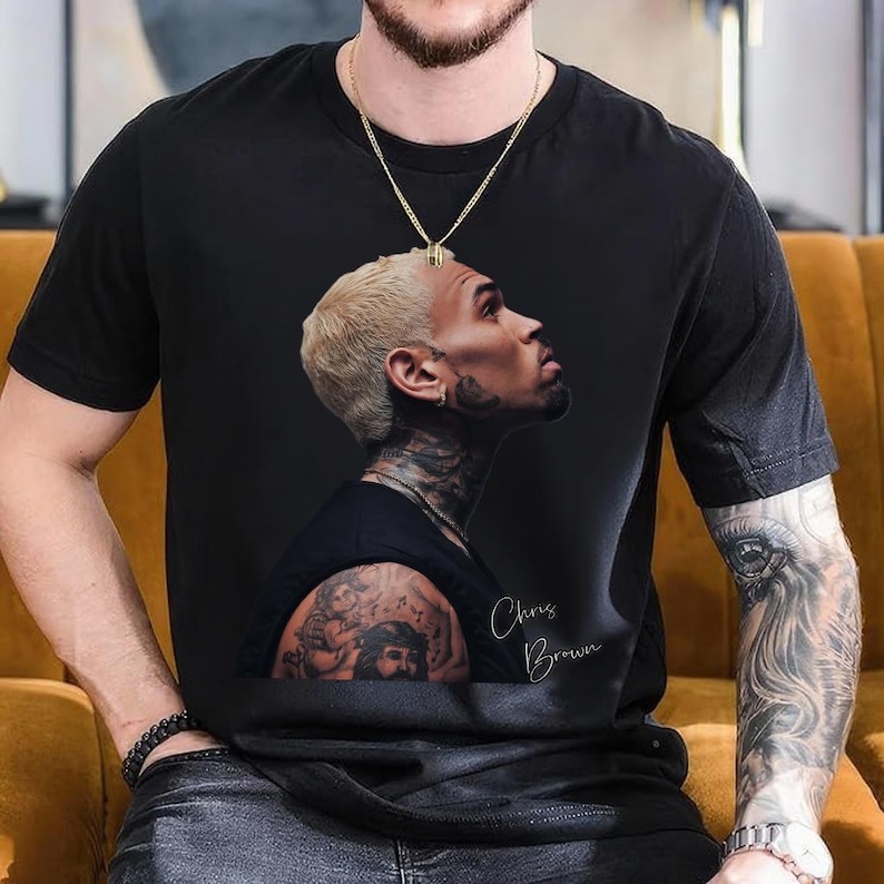 May include: Black t-shirt featuring a side profile portrait of Chris Brown with blonde hair and tattoos. The portrait is in a realistic style, with the name "Chris Brown" written in cursive. A gold chain necklace is visible.