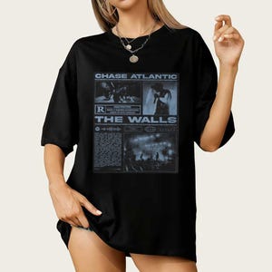 Puede incluir: Camiseta negra con un diseño gráfico azul que incluye el texto "CHASE ATLANTIC" y "THE WALLS". El diseño incorpora imágenes y texto en un estilo de periódico. La camiseta tiene un corte holgado y oversize.
