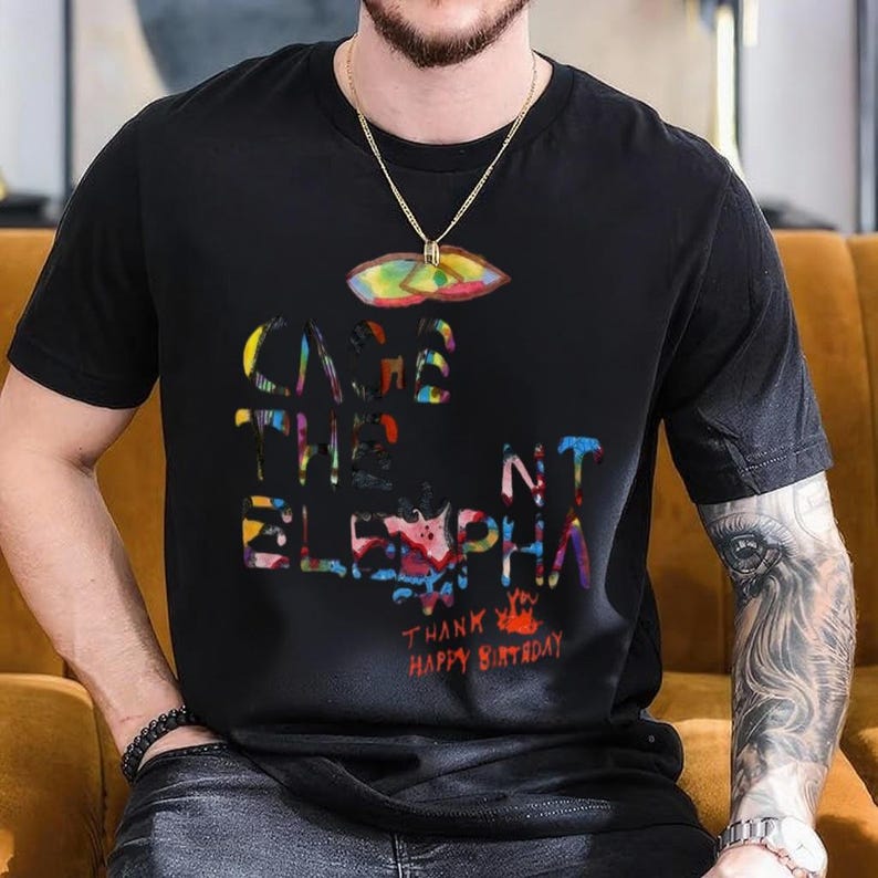 May include: Black t-shirt with a colorful graphic design. The design includes the words "CAGE THE ELEPHANT" in a vibrant, multi-colored font, along with the phrases "THANK YOU" and "HAPPY BIRTHDAY". A gold chain with a pendant is visible.