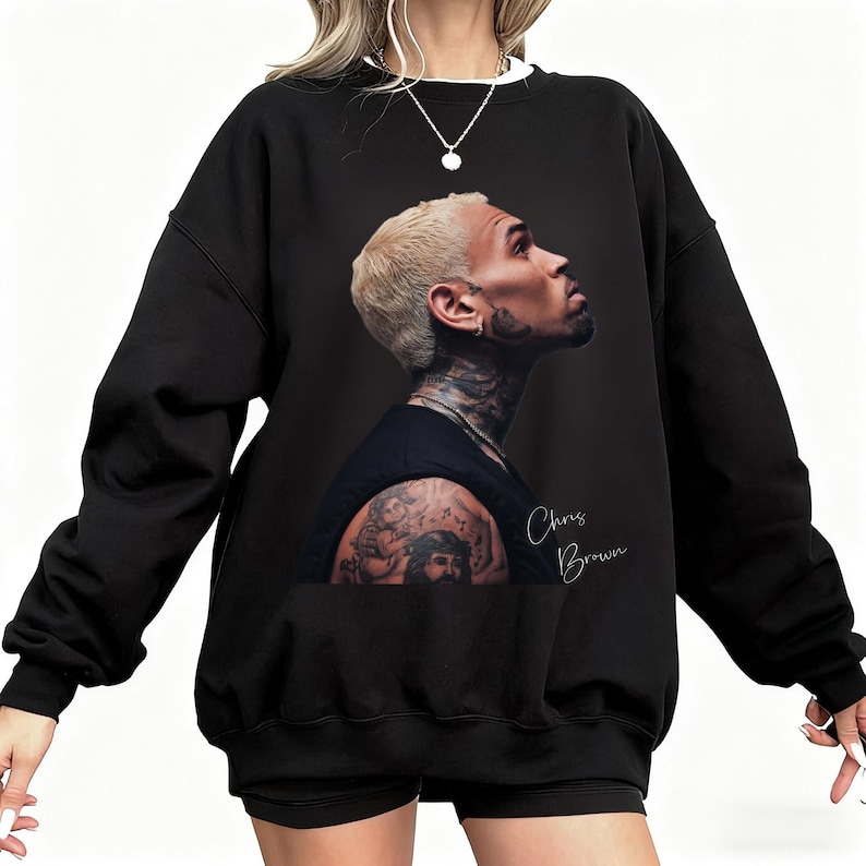 May include: Black sweatshirt featuring a profile image of Chris Brown with blonde hair and tattoos. The name "Chris Brown" is written in cursive on the lower right. The sweatshirt has a white collar.