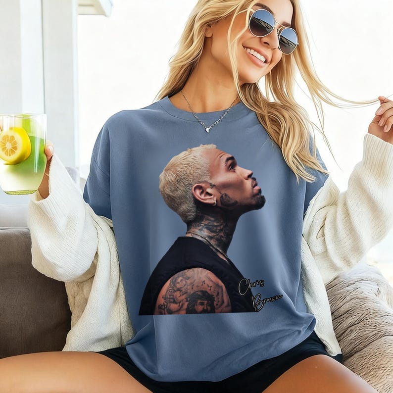 May include: A blue crew neck sweatshirt featuring a portrait of Chris Brown. The image shows a side profile of the singer with tattoos and the name "Chris Brown" in cursive. The sweatshirt is a solid blue color.