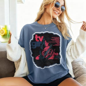 K&ouml;nnte beinhalten: Blau-graues T-Shirt mit einem Grafikdesign. Das Design zeigt die Worte "TV Girl" in Rot und ein schwarz-rotes Bild einer Frau. Das T-Shirt hat eine Vintage-&Auml;sthetik.