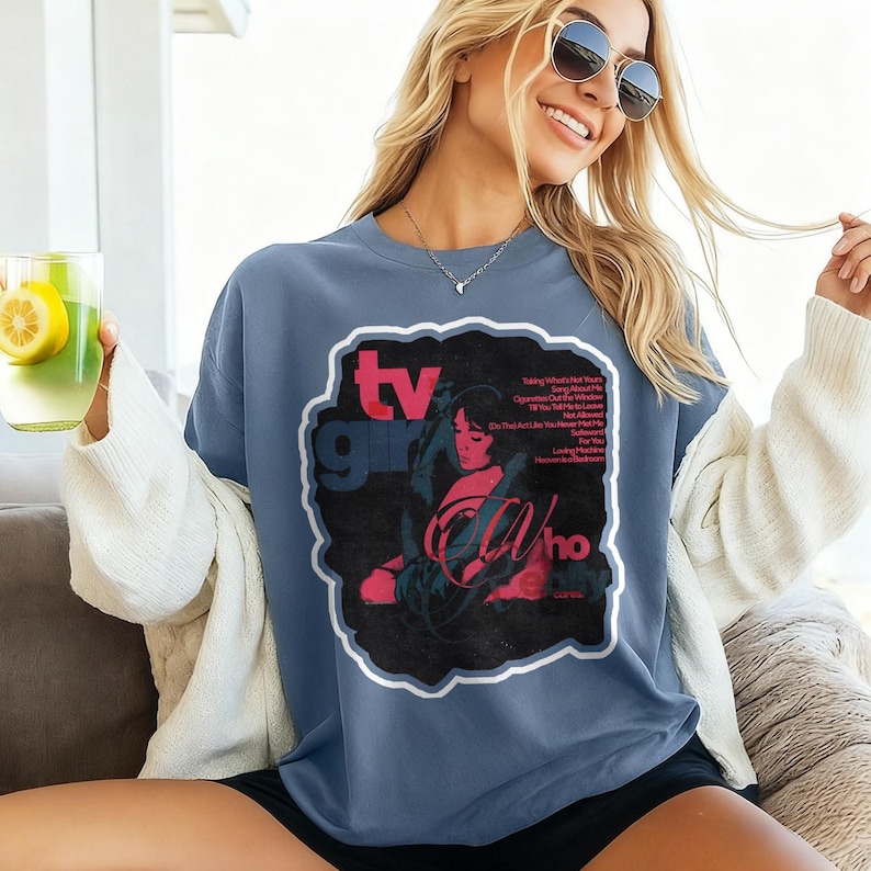 K&ouml;nnte beinhalten: Blau-graues T-Shirt mit einem Grafikdesign. Das Design zeigt die Worte "TV Girl" in Rot und ein schwarz-rotes Bild einer Frau. Das T-Shirt hat eine Vintage-&Auml;sthetik.