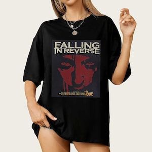Op de afbeelding: Zwart T-shirt met een afbeelding van "FALLING IN REVERSE" boven een rood gezicht. Daaronder staat de tekst "THE POPULAR MONSTOUR". Het shirt heeft korte mouwen en een losse pasvorm.
