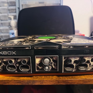 Puede incluir: Consola Xbox negra con diseño de calaveras. La consola tiene un botón de encendido verde y la palabra "XBOX" impresa en la parte delantera. El diseño de calaveras está en la parte superior y frontal.