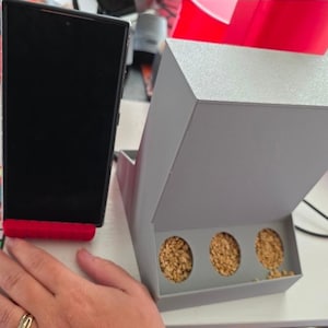 以下が含まれることがあります： 黒いスマートフォンが赤いスタンドに立てかけられています。その隣には、鳥の餌が入った3つの円形の開口部がある灰色の長方形の鳥の餌箱があります。餌箱には蝶番付きの蓋が付いています。