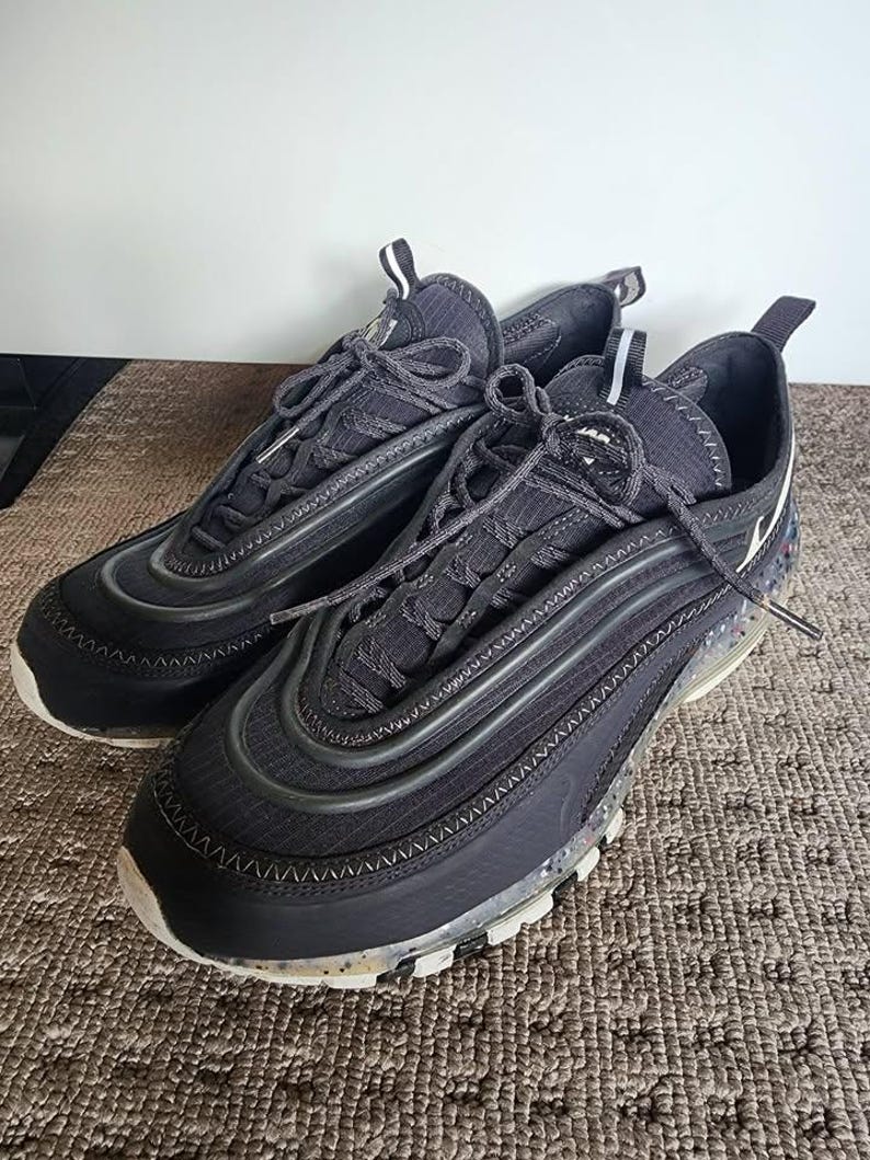 Puede incluir: Un par de zapatillas deportivas negras con un dise&ntilde;o texturizado. Las zapatillas tienen una suela blanca con detalles moteados y cordones negros. Las partes superiores presentan un dise&ntilde;o en capas con una mezcla de materiales y una burbuja de aire visible.