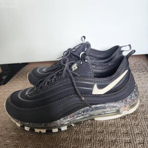 Puede incluir: Par de zapatillas deportivas negras con detalles blancos, incluido un logotipo de Nike blanco. Las suelas son transparentes con motas de colores. Los zapatos est&aacute;n atados y descansan sobre una alfombra marr&oacute;n.