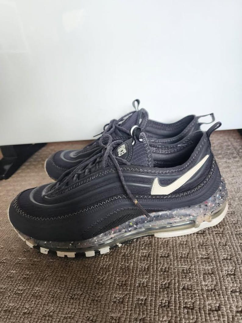 Puede incluir: Par de zapatillas deportivas negras con detalles blancos, incluido un logotipo de Nike blanco. Las suelas son transparentes con motas de colores. Los zapatos est&aacute;n atados y descansan sobre una alfombra marr&oacute;n.