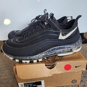 Puede incluir: Zapatillas Nike Air Max Terrascape 97 negras con detalles blancos y suela moteada. Las zapatillas, talla 46, est&aacute;n sobre una caja de cart&oacute;n marr&oacute;n con el logo de Nike e informaci&oacute;n del producto.