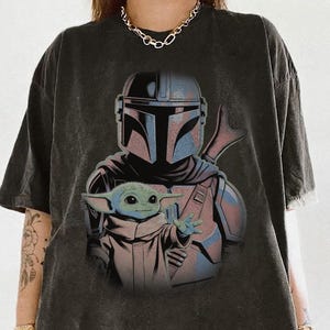 May include: A charcoal gray oversized t-shirt featuring a graphic print of the Mandalorian character wearing a helmet and armor, with the small green creature known as Grogu positioned in the foreground with one hand extended forward.