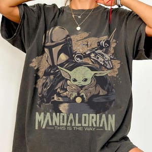 May include: Dark gray t-shirt featuring a graphic of the Mandalorian and Grogu from the Star Wars series. The design includes the text "MANDALORIAN" and "THIS IS THE WAY". The shirt has short sleeves and a relaxed fit.